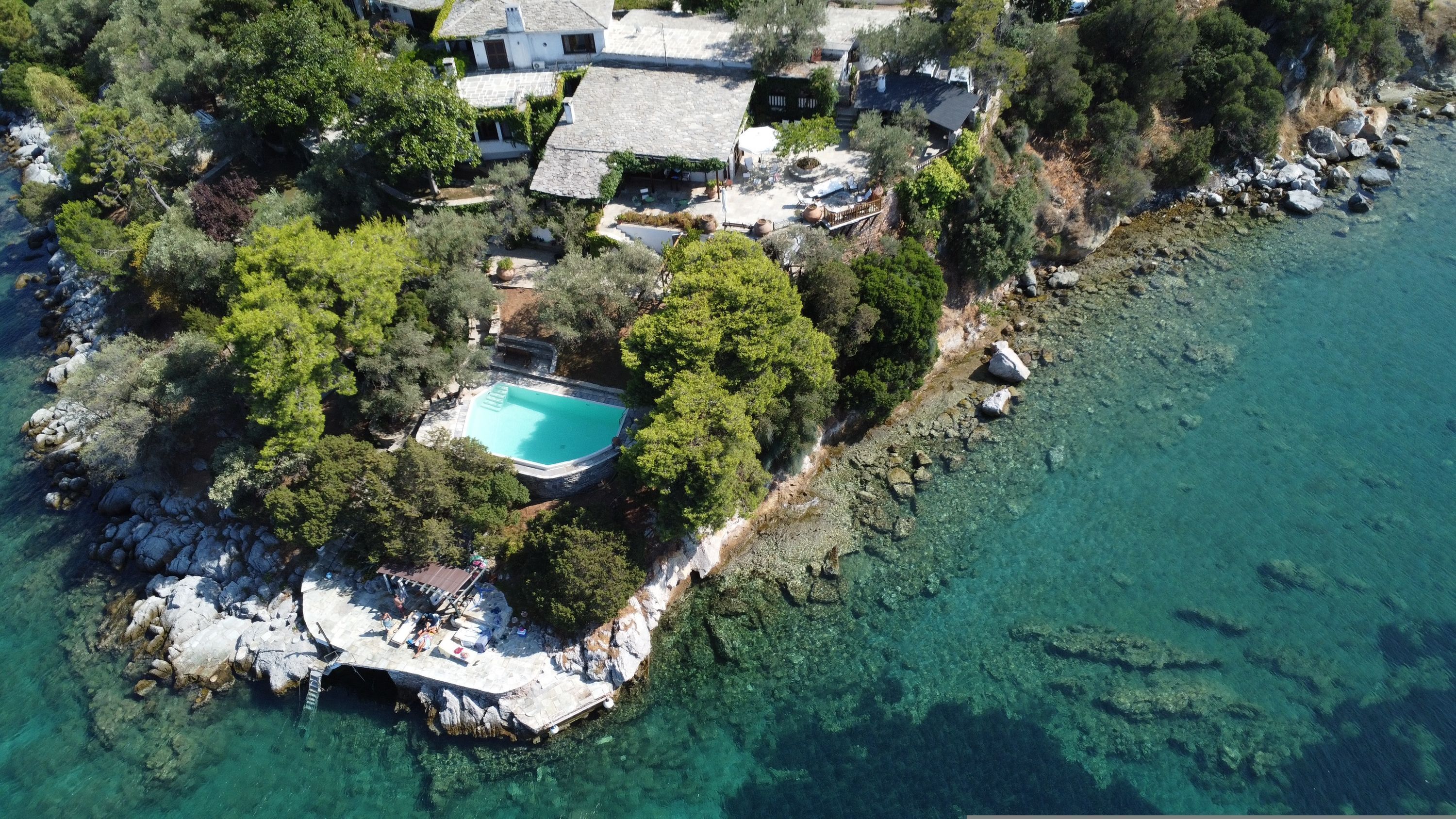 An aerial view of a coastal property with a swimming pool surrounded by trees and a rocky shoreline.