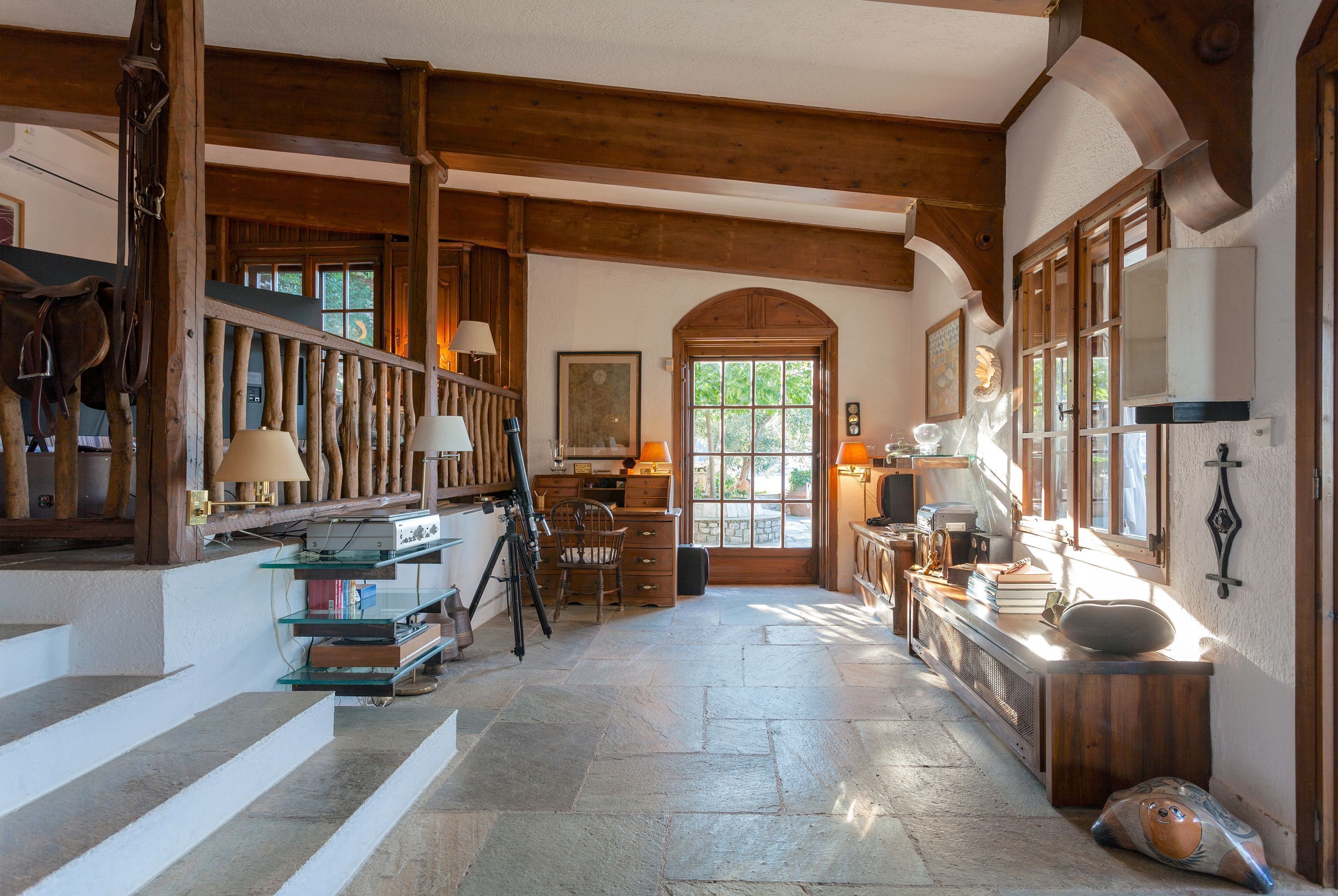 A rustic interior with stone flooring, wooden beams, a staircase, and a desk with books and a lamp.