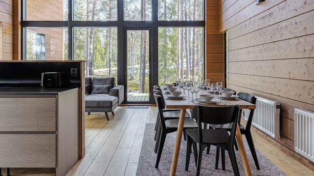Un interior moderno con comedor, cocina y ventanas amplias que dan a un bosque.