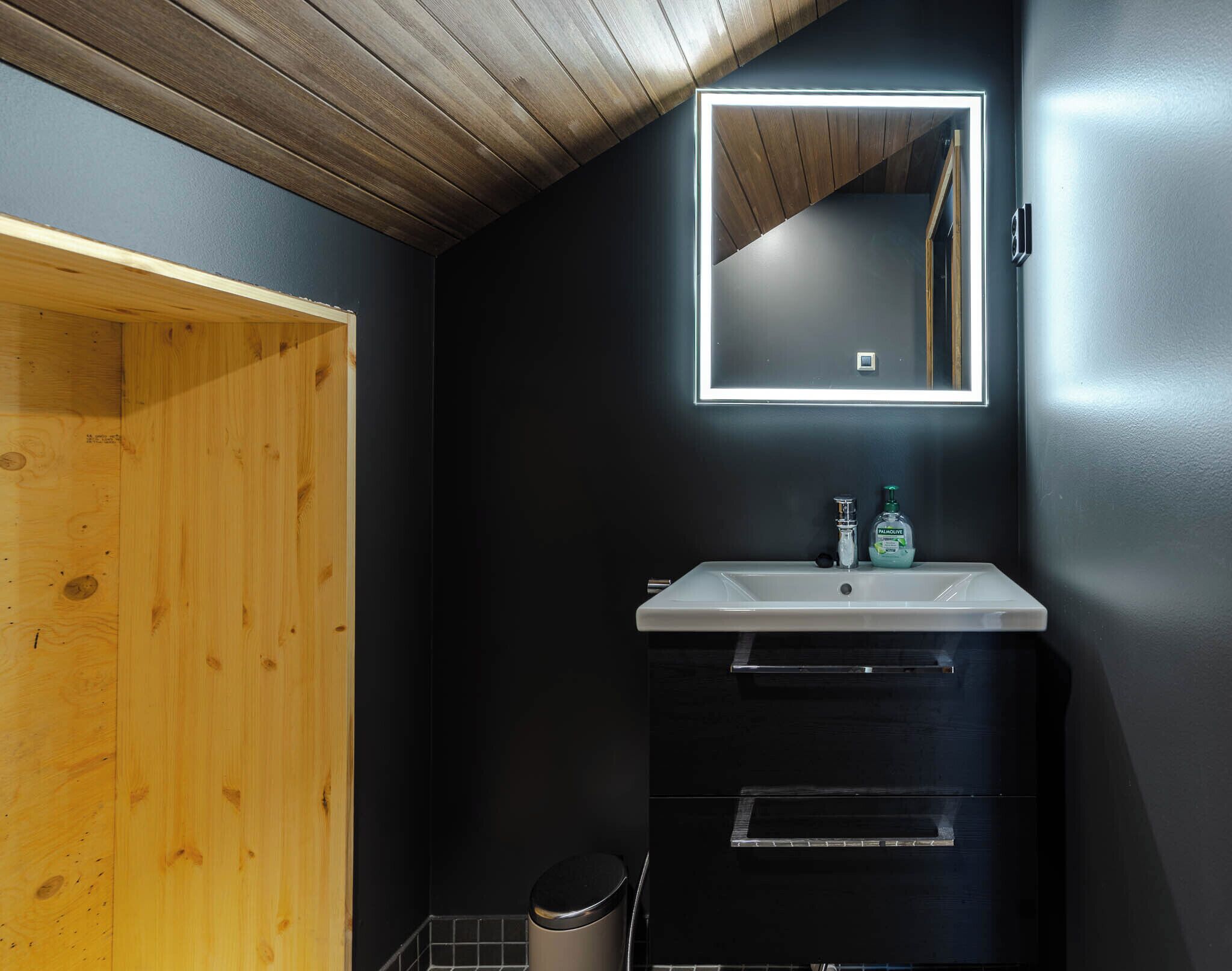 Een badkamer met een houten deur, een wit wastafelmeubel, een spiegel en een prullenbak.