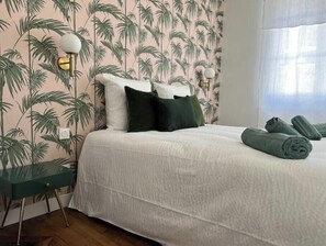 Een slaapkamer met een bed, kussens, een nachtkastje en een muur met een tropisch plantenpatroon.
