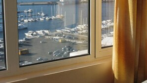 Une vue à travers une fenêtre, avec un rideau, qui révèle un port de plaisance avec des bateaux et une côte lointaine.