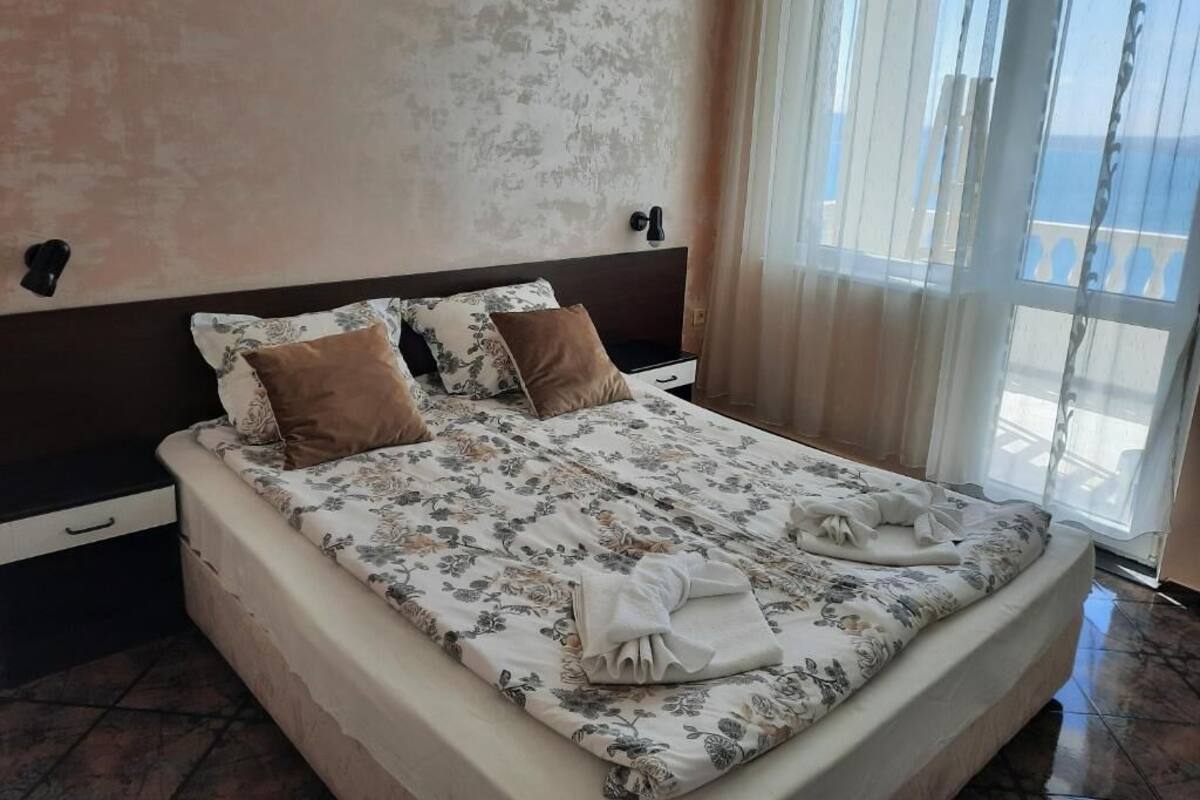 A hotel room with a bed, a nightstand, a window with curtains, and a view of the sea.