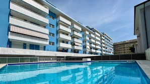 A swimming pool with a tiled deck, surrounded by a multi-story building with balconies. - Poolside apartment with an enchanting terrace (Porto Santa Margherita)