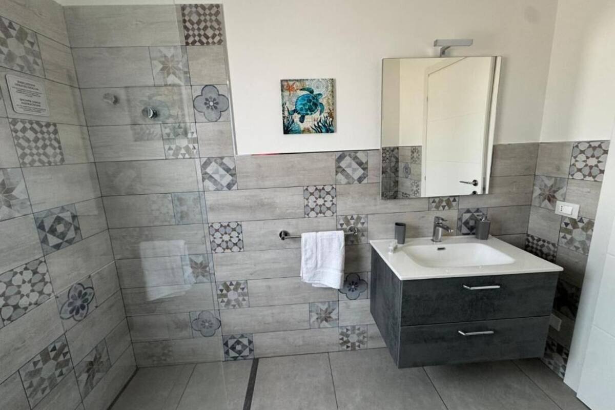 A bathroom with a tiled shower wall, a sink with a mirror, and a towel hanging on the wall.