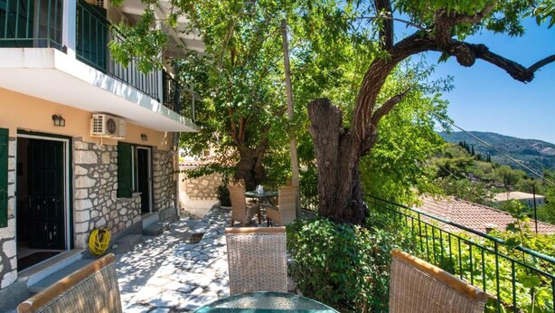 A stone building with a balcony, wicker furniture, and a large tree in a courtyard. - Almondtree House Exanthia - Private Parking (Lefkada)