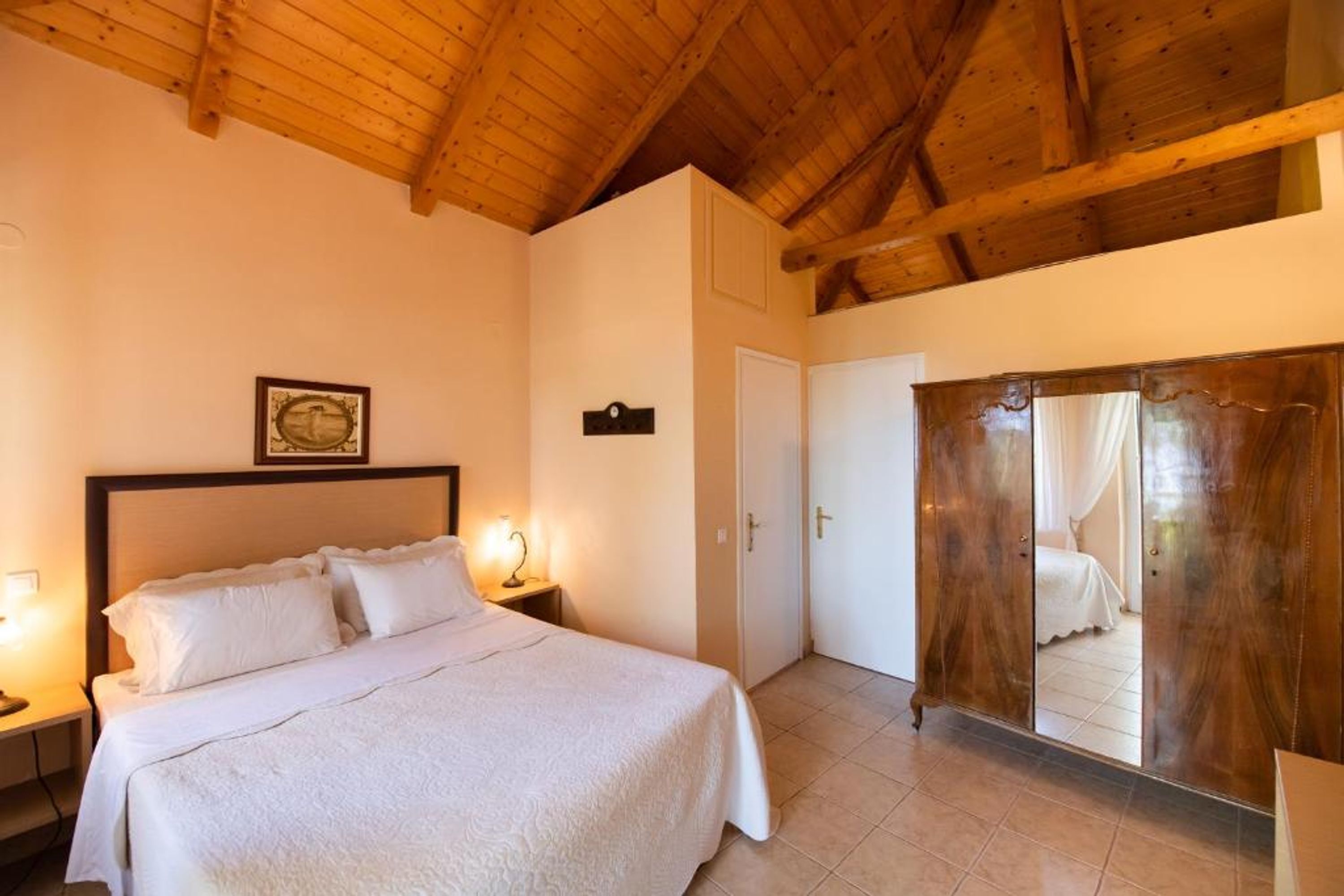 A bedroom with a wooden ceiling, a bed with white bedding, a nightstand with a lamp, a wardrobe with a mirror, and a door leading to another room.