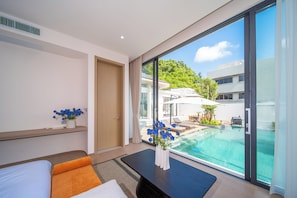A modern hotel room with a large window, a bed, a bedside table with a vase of flowers, and a view of a pool and outdoor seating area.
