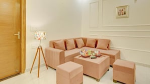A living room with a sofa, ottomans, a floor lamp, and a framed picture on the wall. - Alaya Stays Freesia 3BHK Apartment (Greater Noida)