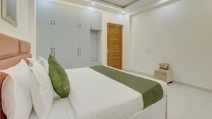 A bedroom with a bed, white wardrobe, wooden door, bedside table, and a framed picture on the wall. - Alaya Stays Freesia 3BHK Apartment (Greater Noida)