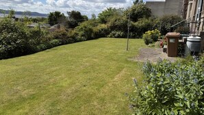 A well-maintained grassy yard with a stone pathway, a metal fence, and a building with a flat roof in the background. - Stylish River View Stay Sleeps 6 (Dundee)