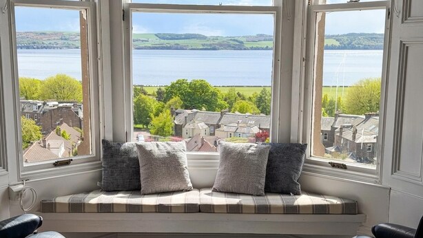 A bay window with a view of a lake and buildings, a bay window seating with cushions, and a black leather chair. - Stylish River View Stay Sleeps 6 (Dundee)