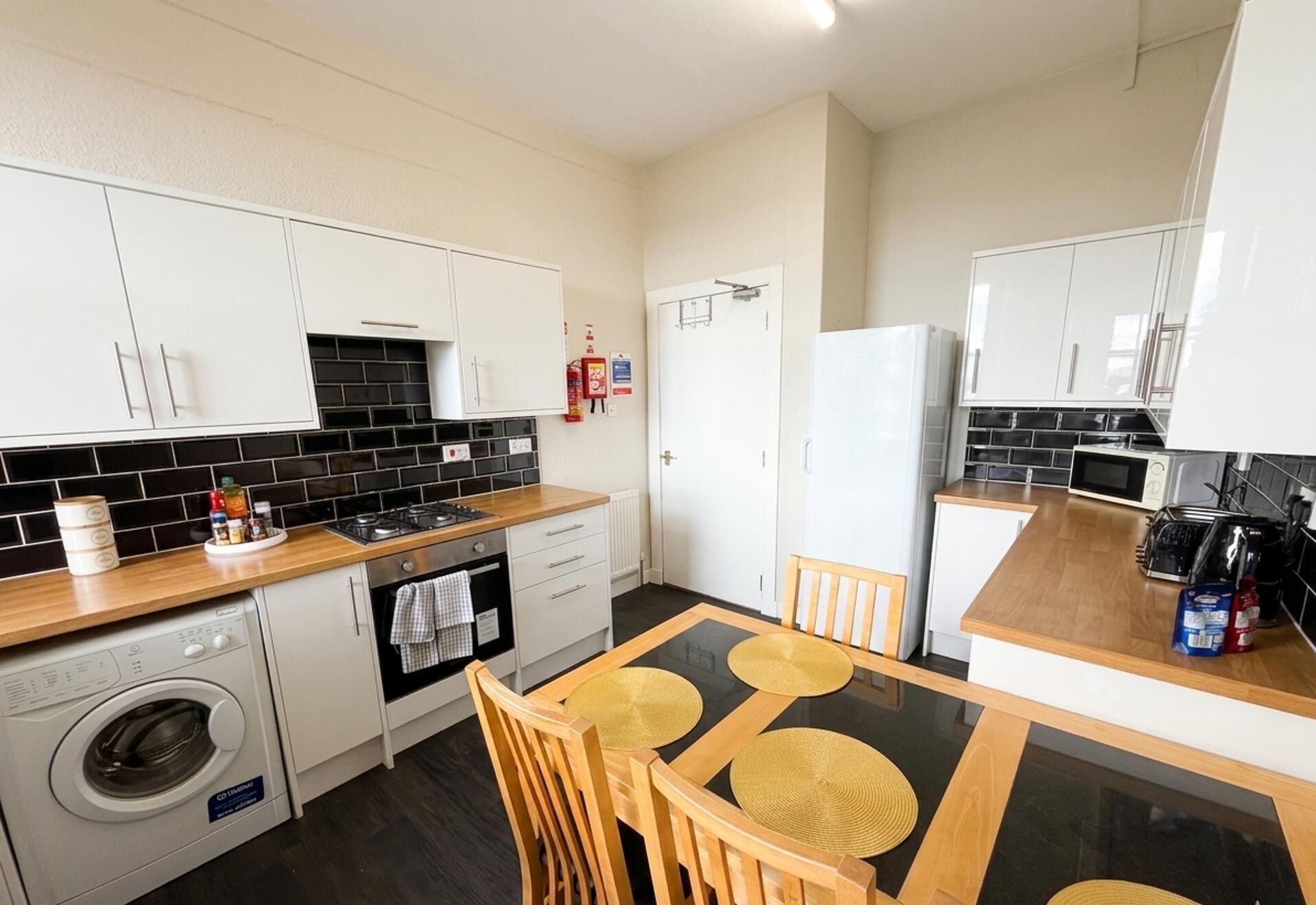 A compact kitchen with white cabinets, a black tiled backsplash, a built-in oven, a microwave, and a small dining area with a table and chairs.