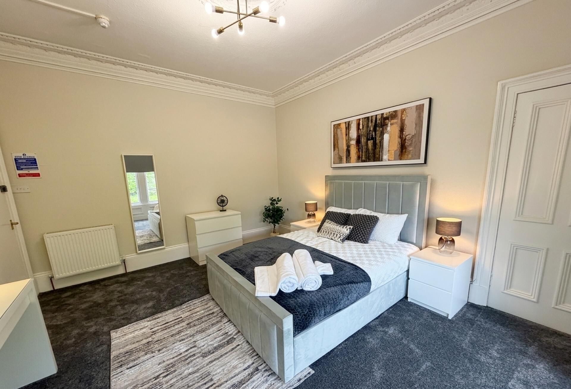 A bedroom with a bed, bedside tables, a mirror, a large windows, and a radiator.