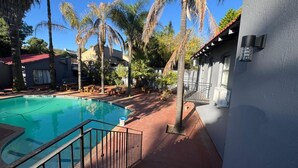 A swimming pool with a red-tiled deck, surrounded by palm trees and a building with a red roof. - Boutique Hotel-Style 8Bed Villa Ferndale (Randburg)