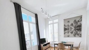 A modern living room with a sofa, a glass coffee table, a dining area with chairs, a TV on a stand, and a large window with curtains. - Le Balcon Notre Dame - 3★ apartment with balcony in the heart of Le Tréport (Le Tréport)