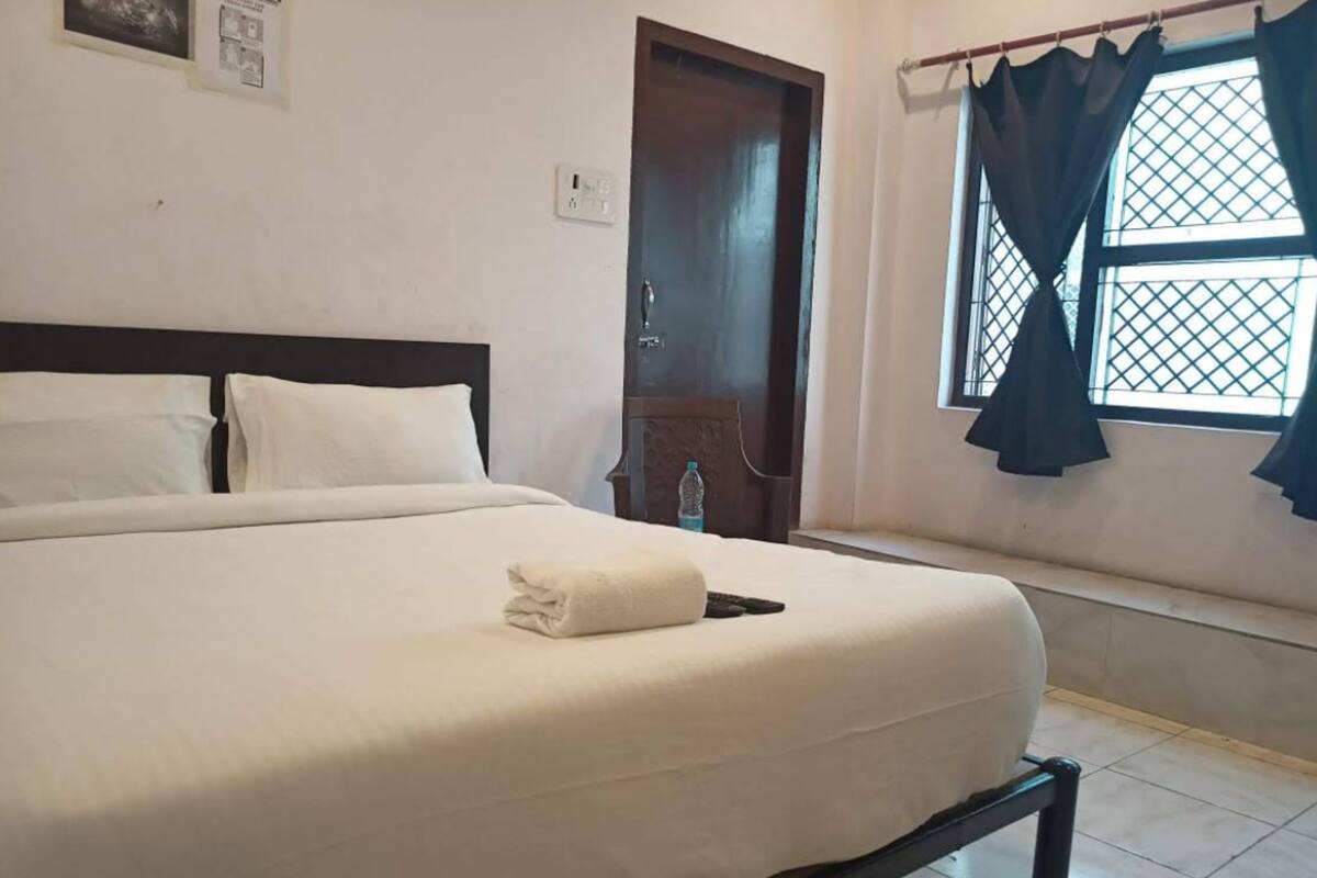 A hotel room with a bed, a window with curtains, a door, and a wall-mounted light switch.