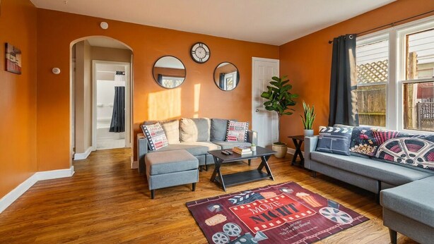 A living room with a sofa, ottoman, coffee table, and a movie-themed rug. There are three wall clocks, a potted plant, and a window with curtains. - Theater Nights, Game Room & Balcony Delights (Rockford)