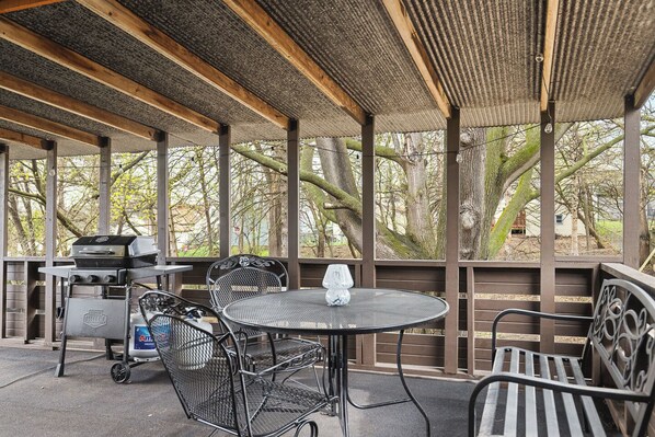 Un patio exterior con una parrilla, sillas de metal, una mesa redonda y un banco.