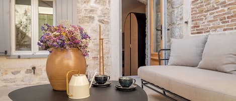 A stone wall, a sofa, a table with a vase and cups, and a window with shutters.