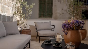 Un espace de détente extérieur accueillant, avec un canapé, un fauteuil, une table avec une théière et un grand vase rempli de fleurs.