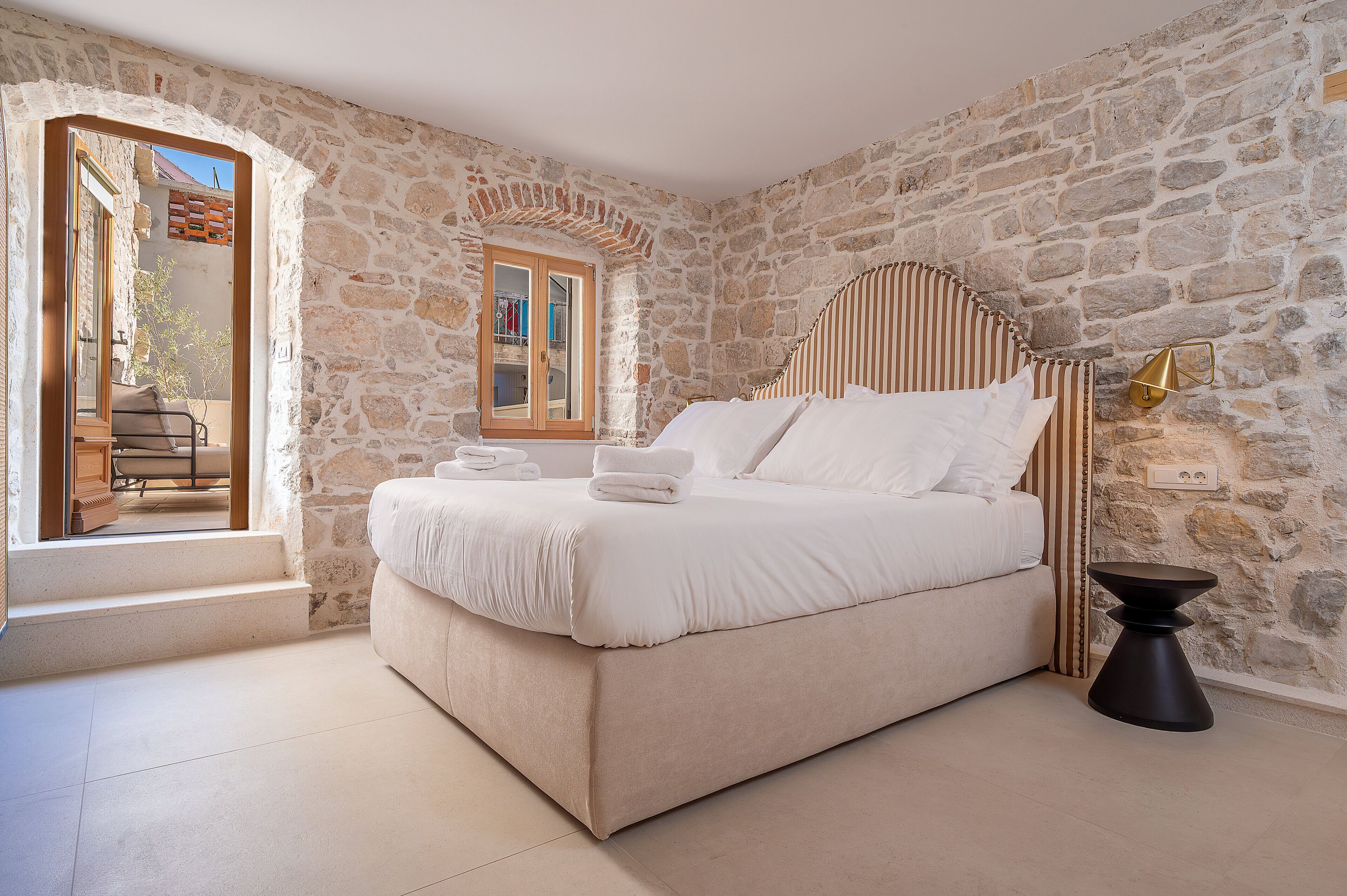 Une chambre avec un mur en pierre, un lit recouvert de draps blancs, une petite table et une fenêtre donnant sur un paysage agréable.