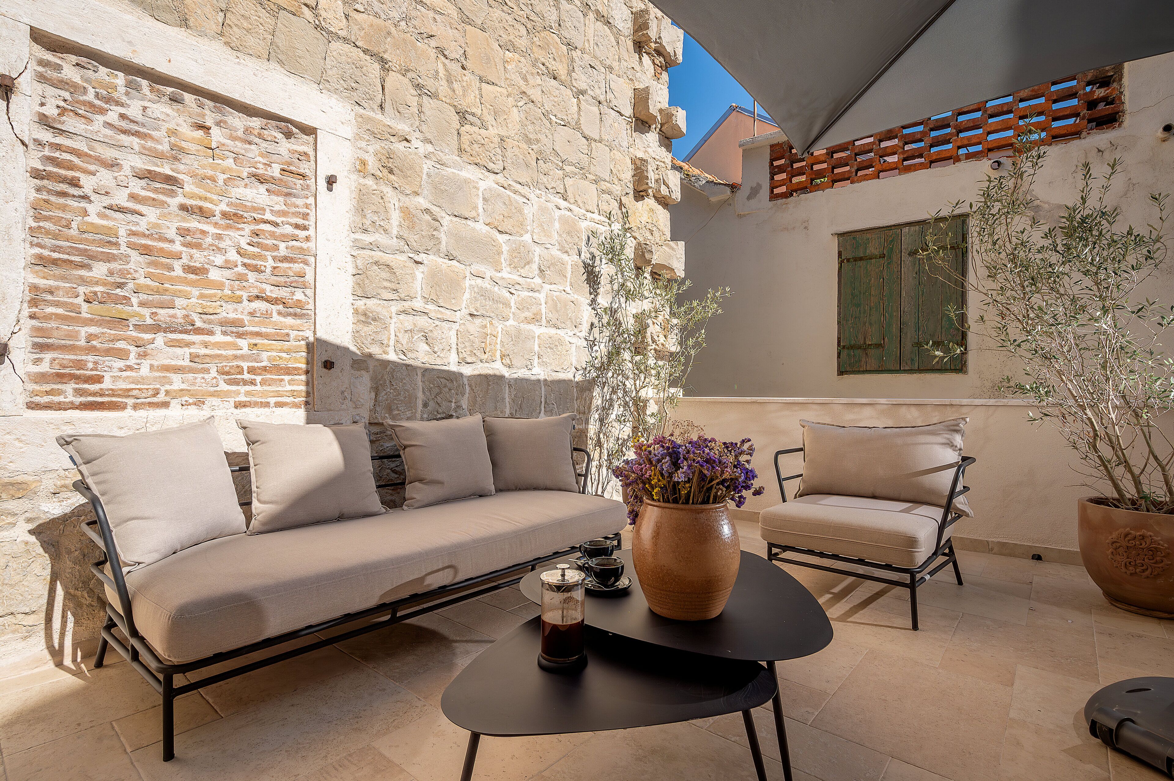 A stone wall, a sofa, a chair, a table, a vase with flowers, and a window with shutters.