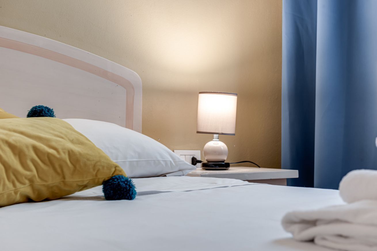 Ein ordentlich bezogenes Bett mit weißen und gelben Kissen, einer Nachttischlampe und einem gefalteten weißen Handtuch auf einem weißen Nachttisch.