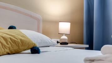 Ein ordentlich bezogenes Bett mit weißen und gelben Kissen, einer Nachttischlampe und einem gefalteten weißen Handtuch auf einem weißen Nachttisch.