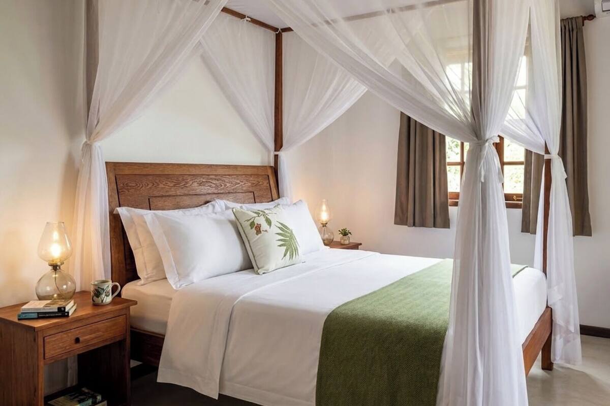 Uma cama com dossel e cortinas, roupa de cama branca e um cobertor verde, mesas de cabeceira de madeira com abajures e uma janela com cortinas.