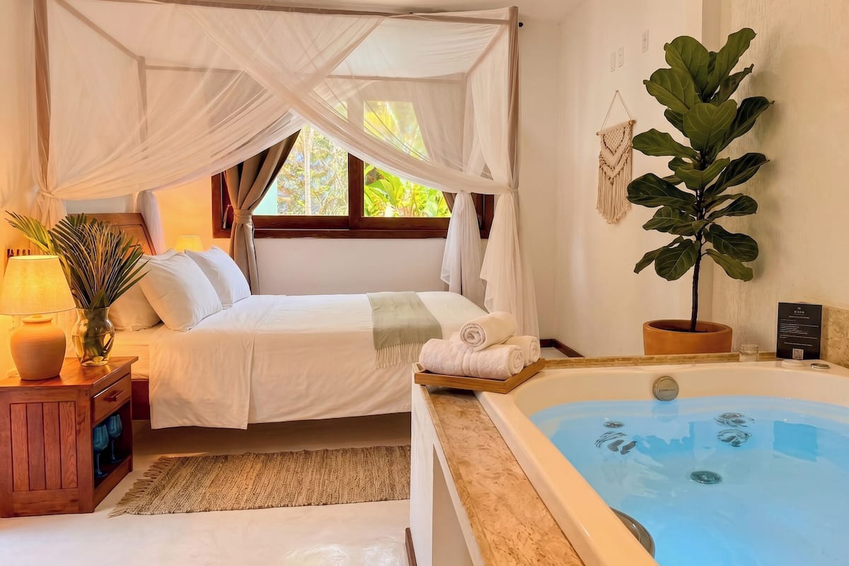 Quarto com cama de dossel, mesa de cabeceira com abajur, planta em vaso e banheira de hidromassagem.