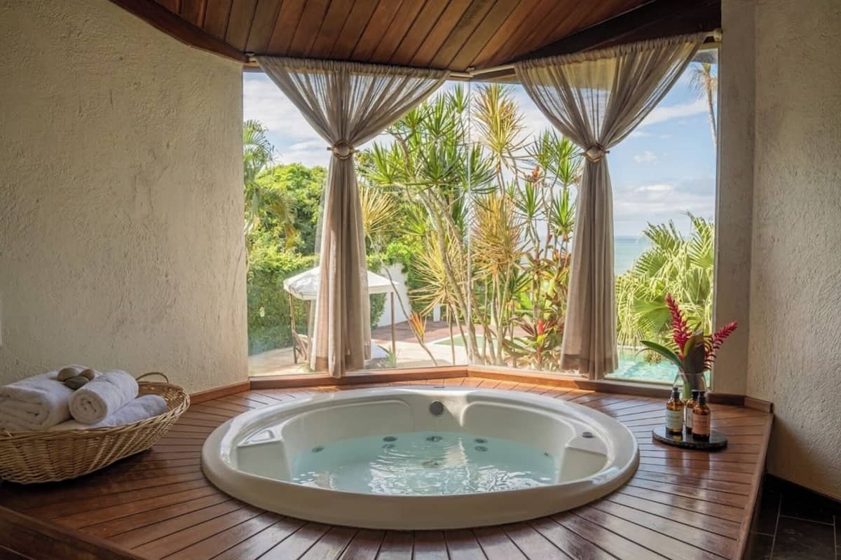 Uma banheira de hidromassagem em área de deck de madeira, com vista para plantas tropicais e uma piscina.