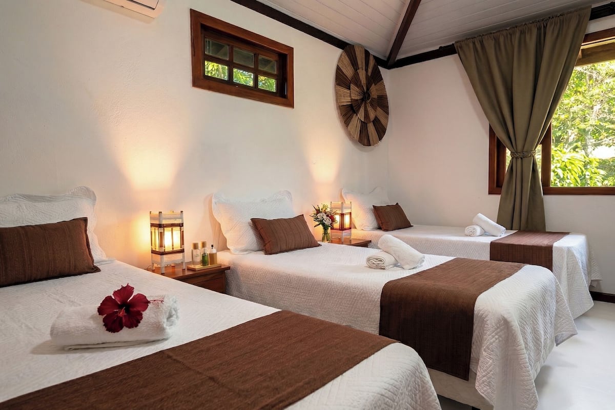 Quarto de hotel com duas camas, um relógio de parede de madeira, uma janela com cortinas e uma mesinha de cabeceira com luminária e artigos de higiene pessoal.