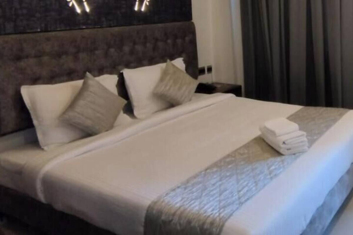 Una habitación de hotel con una cama grande, cabecero, mesita de noche y ventana con cortinas.