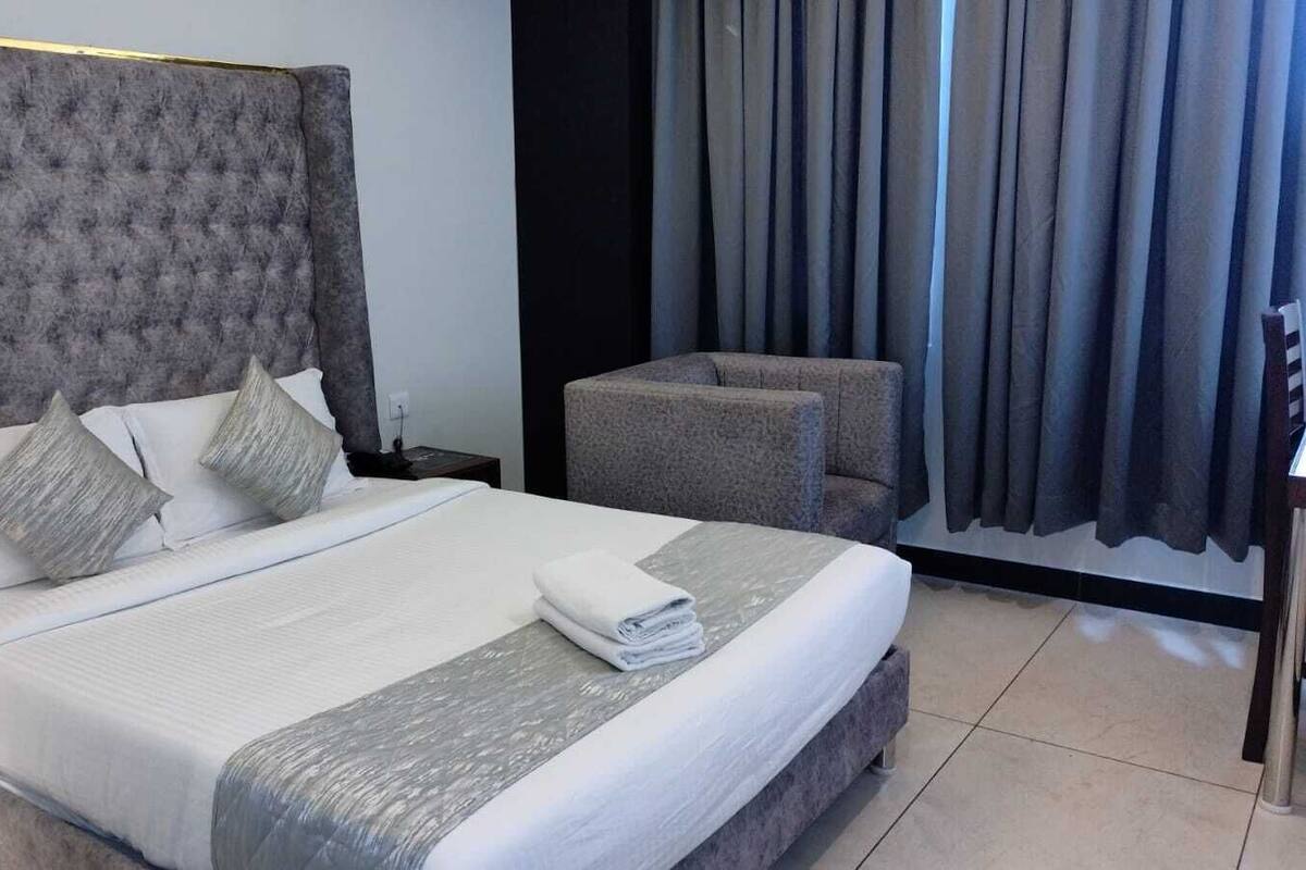Una cama bien hecha con almohadas, un sillón tapizado de color gris y una ventana con cortinas.