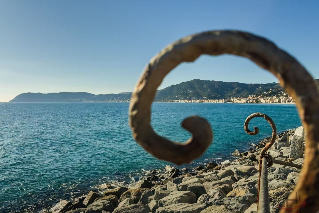 A coastal view with a metal spiral structure in the foreground, rocky shoreline, and distant buildings.
