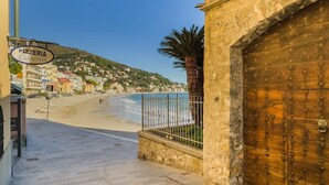 Une rue côtière avec une porte en bois, une enseigne indiquant « Pizzeria La Lanterna », et une plage en arrière-plan.