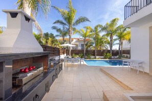 Eine Terrasss mit Grill, Pool und Palmen.
