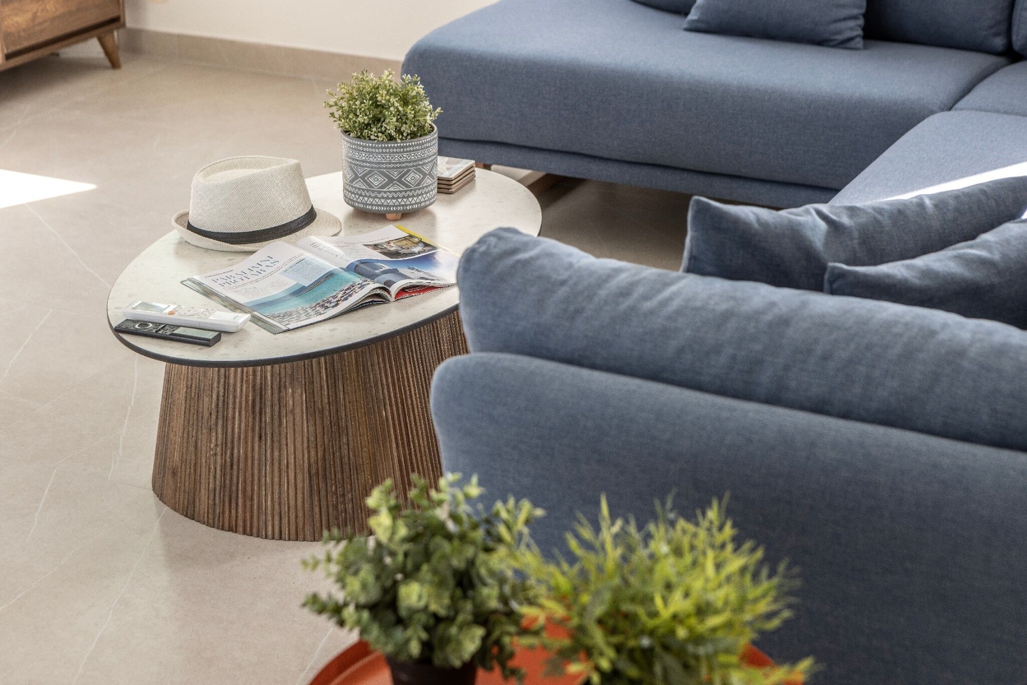 A modern living room with a blue sofa, a round wooden coffee table with magazines and a hat, and a potted plant.