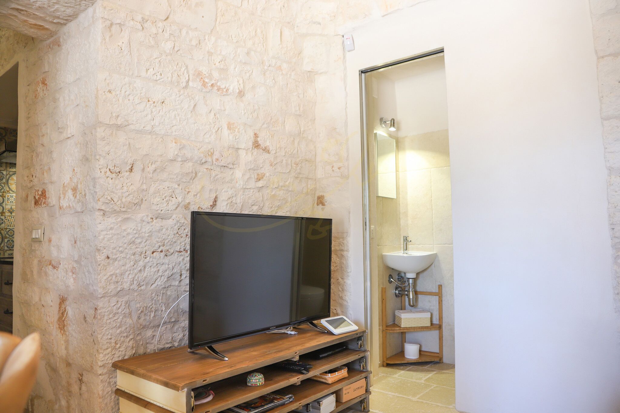 A room with a stone wall, a flat-screen TV on a wooden stand, a small white shelf, and a bathroom with a sink and mirror.