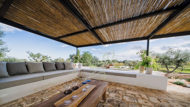 An outdoor seating area with a stone floor, a wooden table with wine glasses, a cushioned bench, and a view of the landscape. - TD Trullo Lumina Boutique Luxury Trulli With Pool (Ostuni)