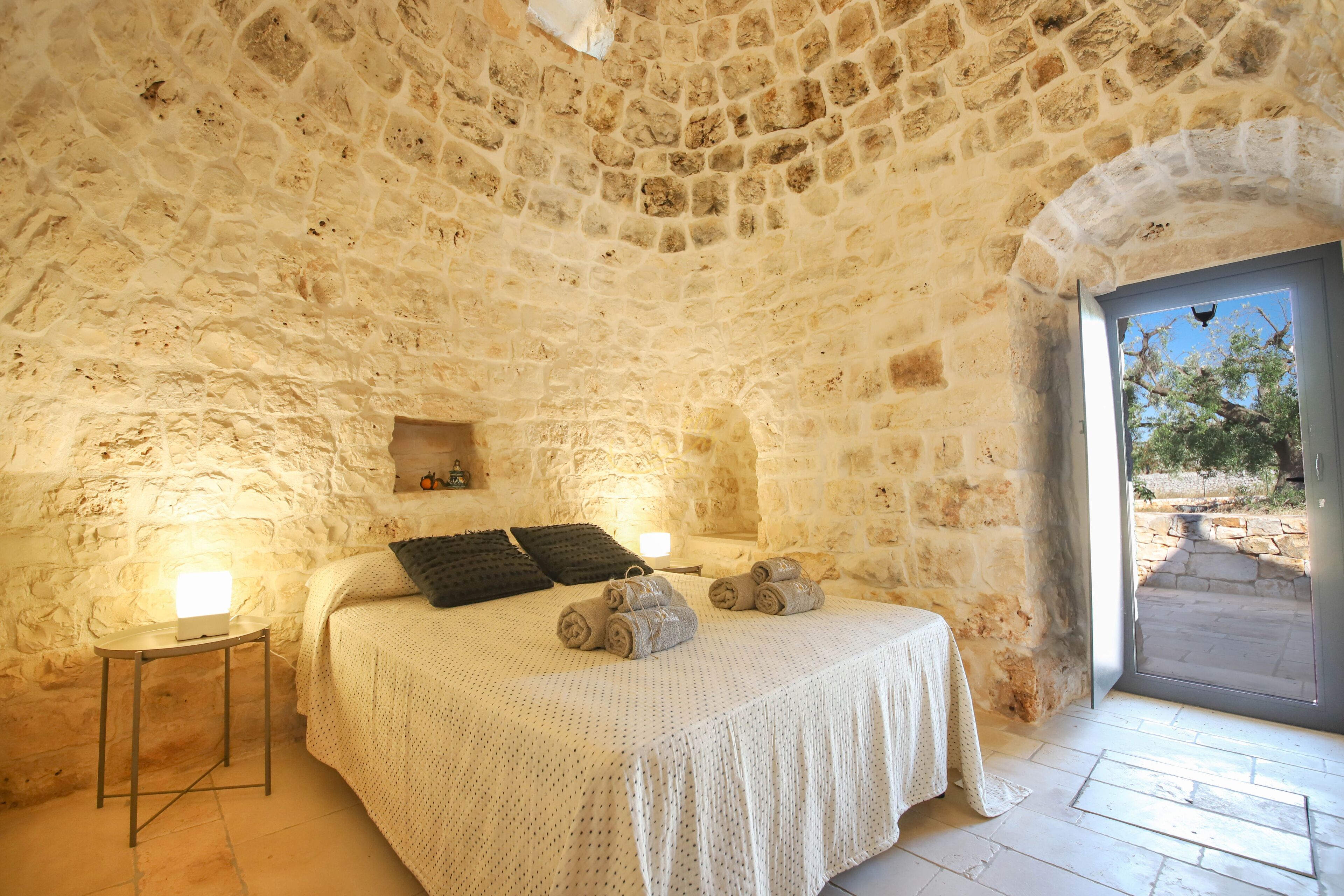 Un dormitorio con paredes de piedra, una cama, lámparas de noche y vistas a través de un puerta abierta.