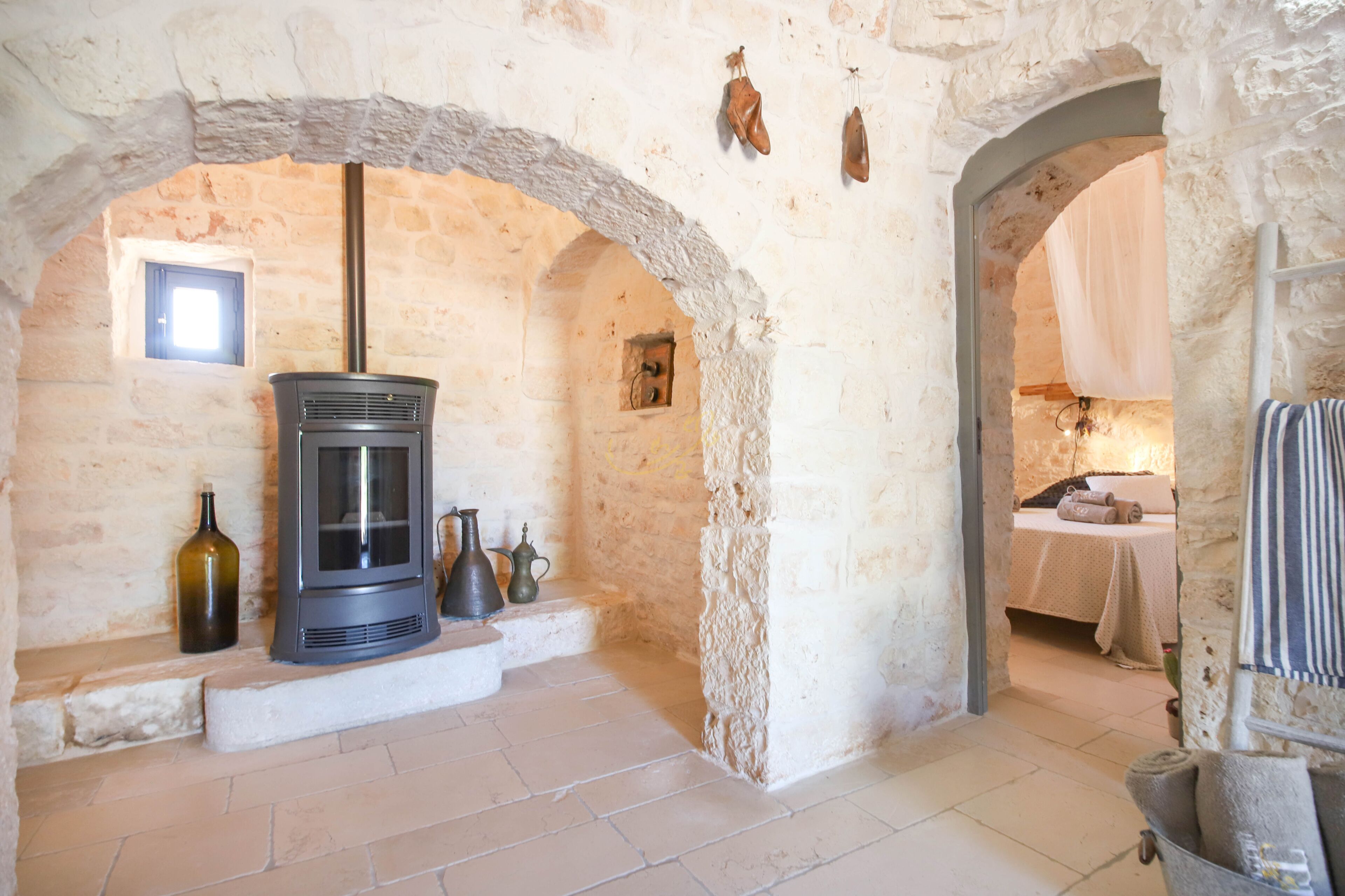 A stone-walled room with a fireplace, a small table with a bottle and two pitchers, and a bed with a white canopy.