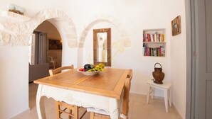 A dining area with a wooden table, chairs, a fruit bowl, a bookshelf, and a small table with a pot. - TD Trullo Buenavista Stunning Valley Views W/pool (Ostuni)