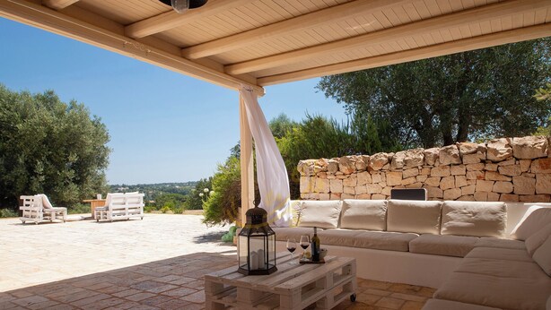 An outdoor seating area with a stone wall, a white sofa, and a wooden table with a lantern. - TD Trullo Buenavista Stunning Valley Views W/pool (Ostuni)