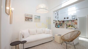 A modern living room with a white sofa, a wicker chair, and a small side table. There is a kitchen area with a countertop and a radiator. - TD Casa Altomare w/ Terrace Over Historic Center (Monopoli)