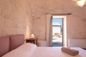 A bedroom with a stone wall, a bed with a headboard, a nightstand, a lamp, and a view of the outside. - TD Trulli Lorusso Design Luxury Trulli With Pool (Castellana Grotte)