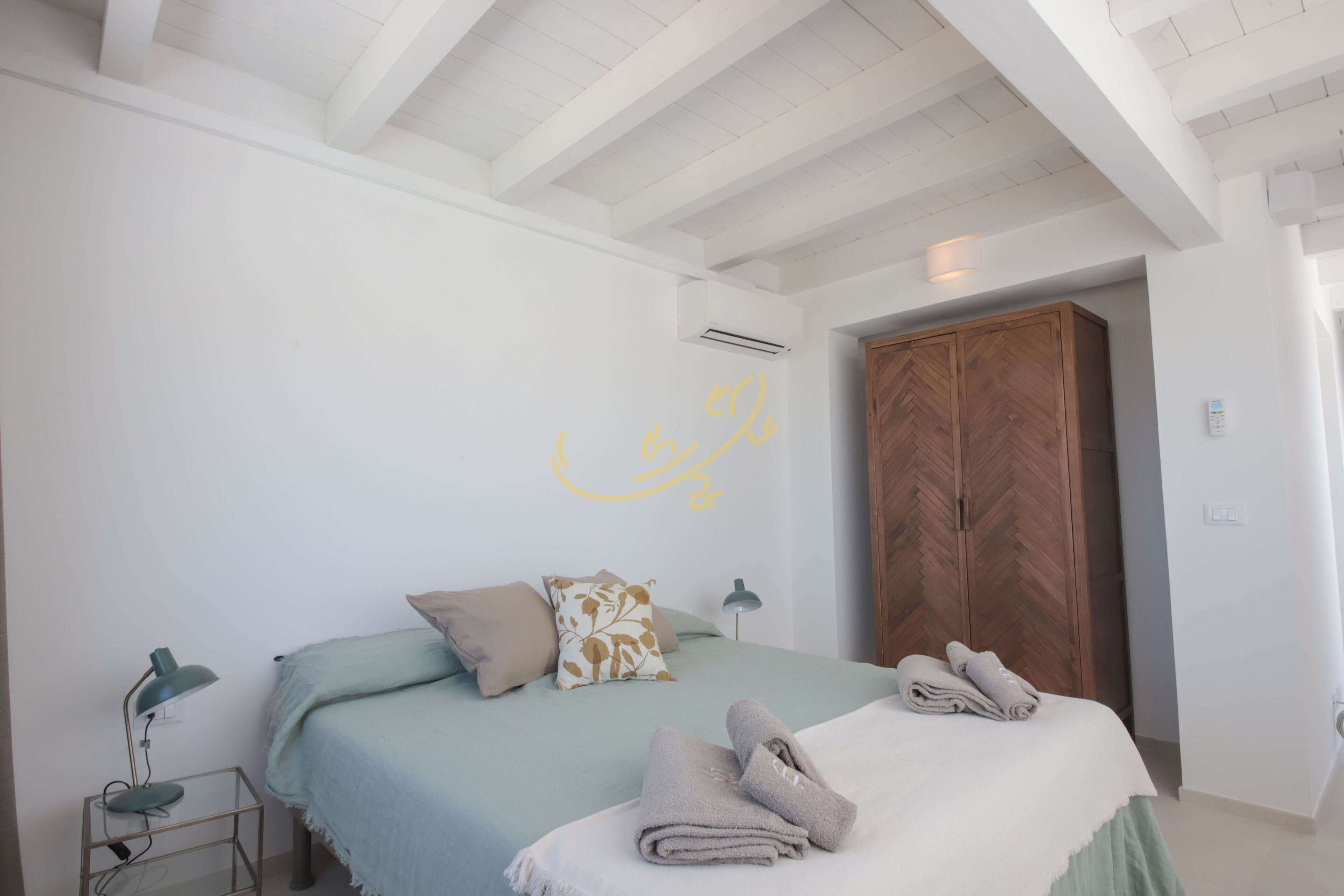 A bedroom with a bed, pillows, a wooden wardrobe, and a wall with a yellow design.