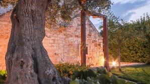 A stone building with a large tree in front, a metal structure, and a grassy area with flowers. - TD Torretta San Procopio Stone House With Pool (Castellana Grotte)