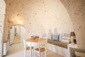 A stone-walled room with a round wooden table, white chairs, a bench with cushions, and a lamp on a side table.